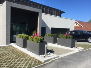 Bildet viser en moderne bygningens inngangsparti med to rektangulære, grå plantekasser på hver side av trappen. Plantekassene er fylt med pent beskårne grønne busker, og hver kasse har to norske flagg som stolt vaier i vinden. Bygningen har en mørk inngangsdør som kontrasterer mot den lyse eksteriørveggen. Foran på bildet er det en økoparkering med gress som vokser gjennom rutenettet på bakken, og en luksusbil parkert til høyre, noe som gir en følelse av ordnet eleganse og nasjonal stolthet.